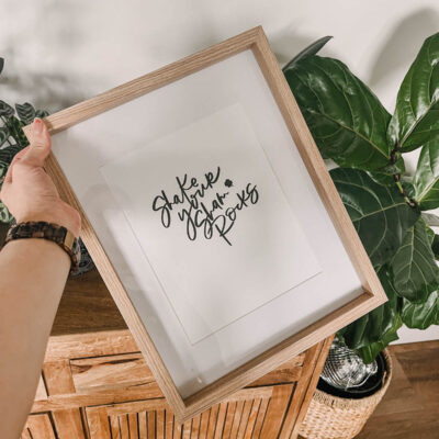 shake your shamrocks hand lettered art in a frame, held in front of plants