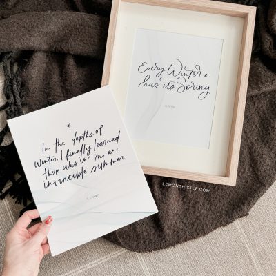Free printable winter quotes shown printed on cardstock, one in wooden frame on cozy blanket